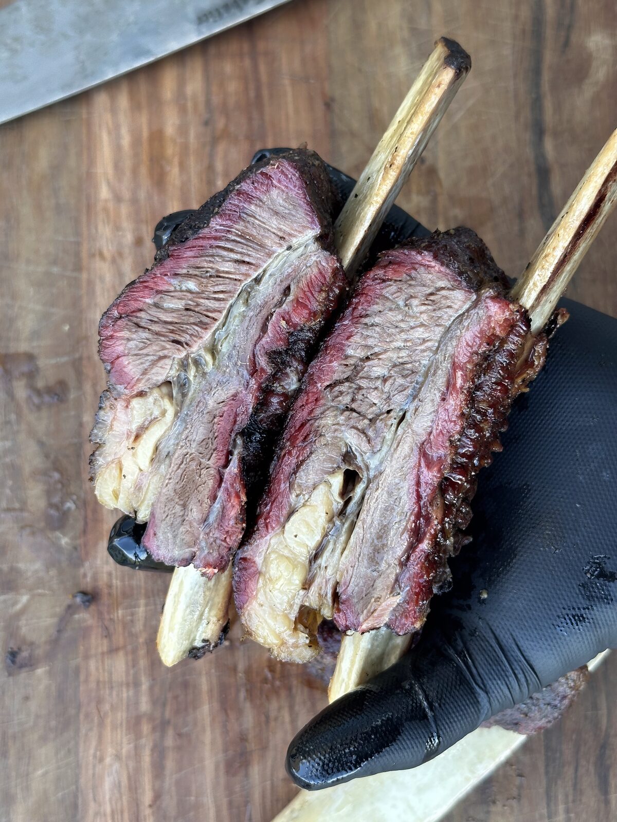Cross-section of smoked beef short ribs held in a gloved hand, showing a pink smoke ring under dark bark